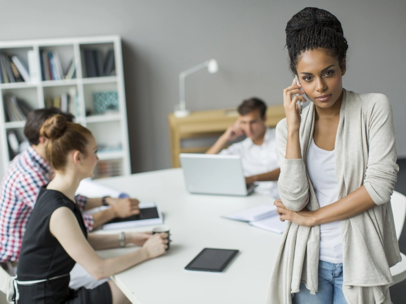 A woman stands apart from a meeting table with a phone to her ear while colleagues keep talking